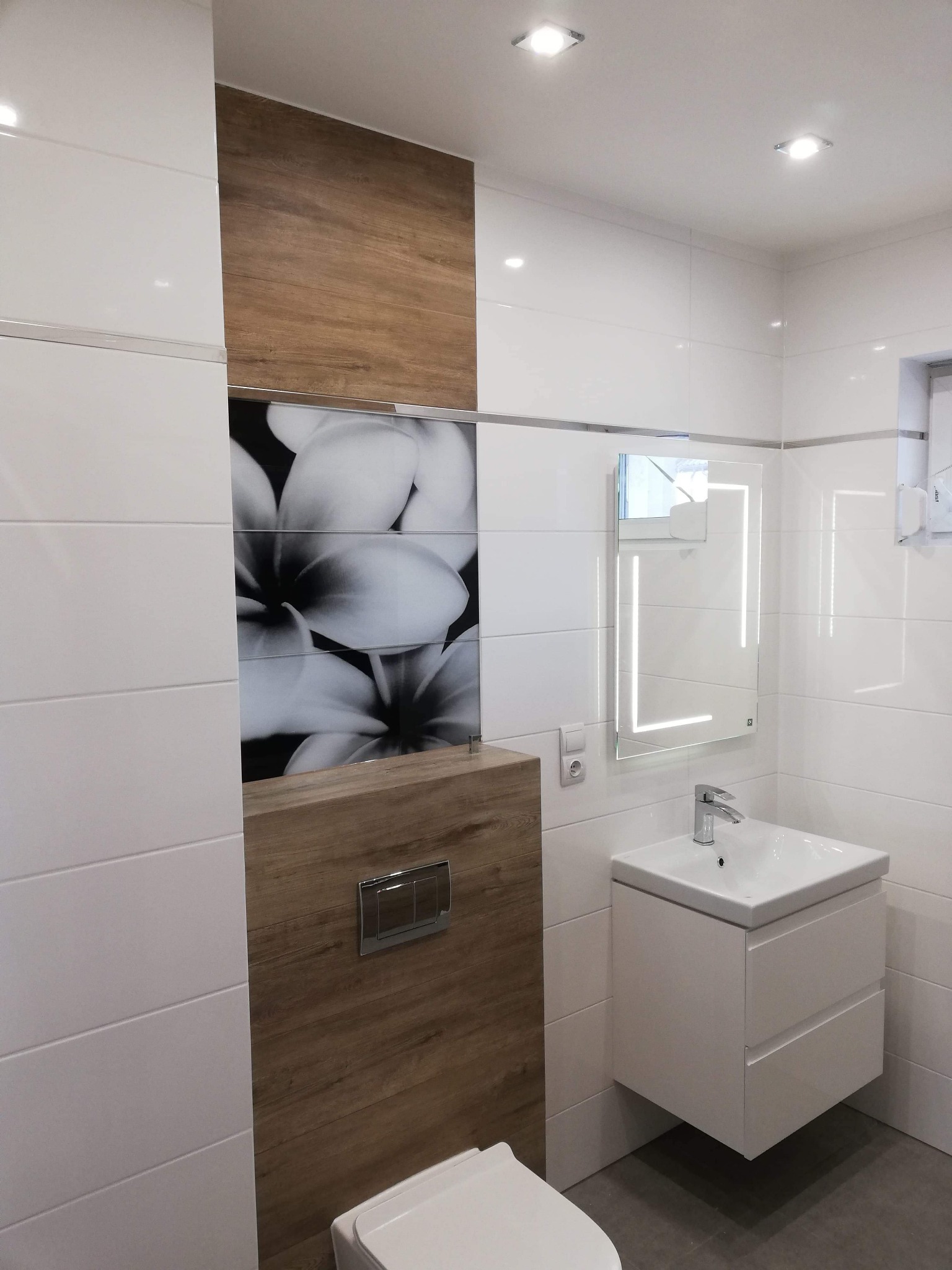 Nowoczesna łazienka z białymi płytkami, akcentem drewna i kwiatowym motywem, podświetlane lustro.