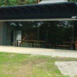 Nowoczesny budynek z cegły i szkła z płaskim dachem, otoczony zielenią, z widocznymi ławkami wewnątrz.