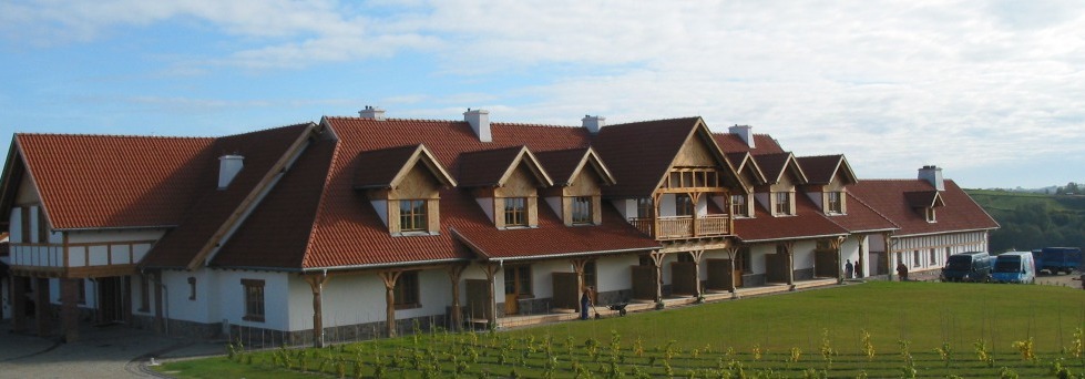 Rozległy budynek murowany z czerwonym dachem, elementami drewnianymi i białymi ścianami, otoczony zielonym trawnikiem i młodymi roślinami, na tle błękitnego nieba.