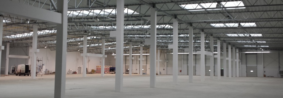 Wnętrze nowo wybudowanej hali z białymi, stalowymi słupami nośnymi, widoczne świetliki w dachu, częściowo wyposażona, z zaparkowanym vanem i rusztowaniem w tle.
