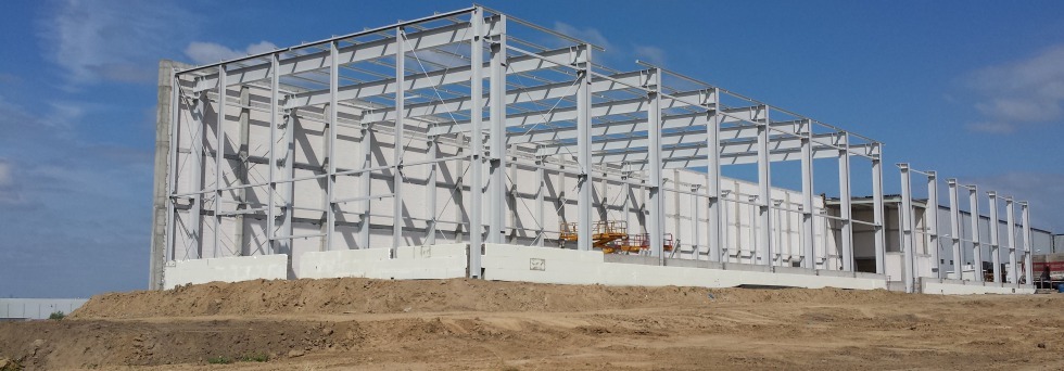 Konstrukcja stalowa hali w trakcie budowy, widoczne stalowe ramy i betonowe fundamenty, na drugim planie podnośnik koszowy