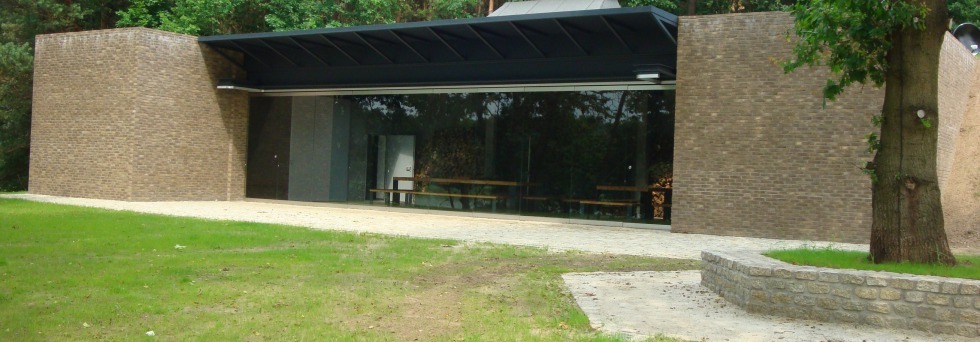 Nowoczesny budynek z cegły i szkła z płaskim dachem, otoczony zielenią, z widocznymi ławkami wewnątrz.