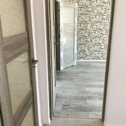 JustDevelopment - Wrocław: Widok na wnętrze po montażu drzwi wewnętrznych z drewnianymi ościeżnicami i przeszklonymi skrzydłami, jedna z nich jeszcze w folii ochronnej, na podłodze panele imitujące drewno, ściana...