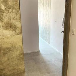 JustDevelopment - Korytarz w mieszkaniu z szarą podłogą z płytek, ściany w odcieniach bieli i beżu, widoczny domofon i włącznik światła, drzwi z drewnianą ościeżnicą.