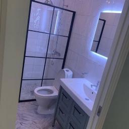 JustDevelopment - Nowoczesna łazienka z kabiną prysznicową typu walk-in z czarnymi szprosami, podświetlanym lustrem i umywalką z szafką w kolorze szarozielonym.