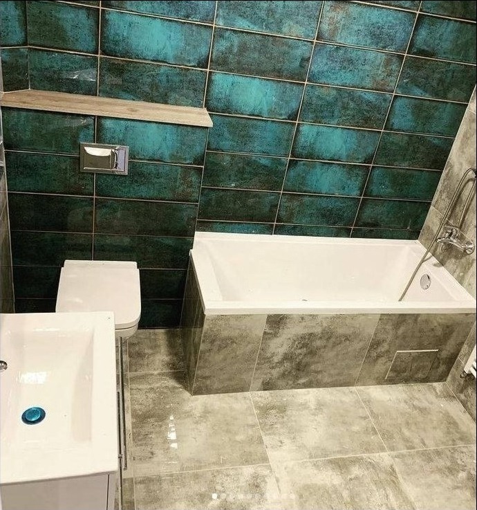 Wykończona łazienka z turkusowymi płytkami ściennymi ułożonymi w cegiełkę, białą wanną obudowaną szarymi płytami imitującymi beton, umywalką z szafką i toaletą.