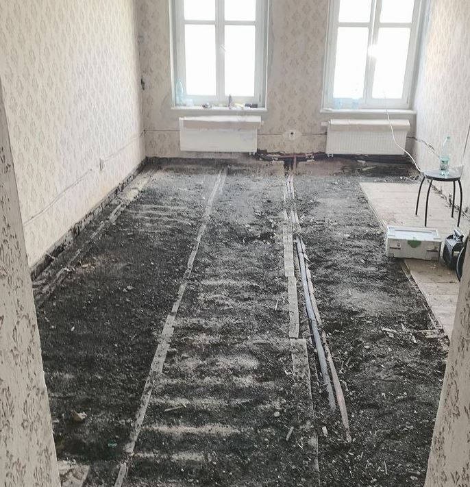 Pokój w trakcie prac przygotowawczych do ułożenia podłogi, widoczne ślady po usuniętej podłodze i listwy przypodłogowe, dwa okna z białymi grzejnikami pod spodem, ściany w jasnej tapecie w drobny...