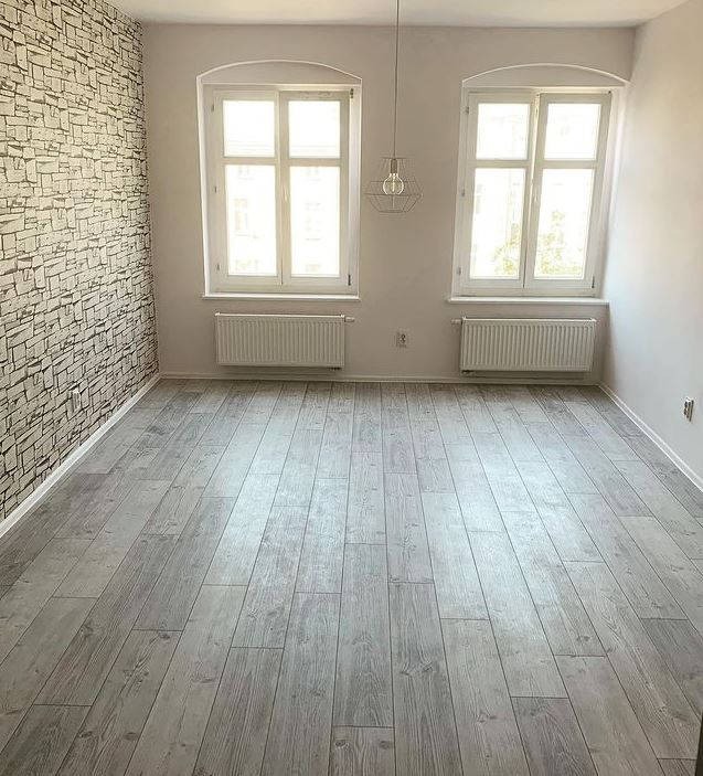 Wnętrze pokoju z szarą podłogą imitującą drewno, białe okna z grzejnikami, ściana z tapetą imitującą kamień i nowoczesna lampa druciana.