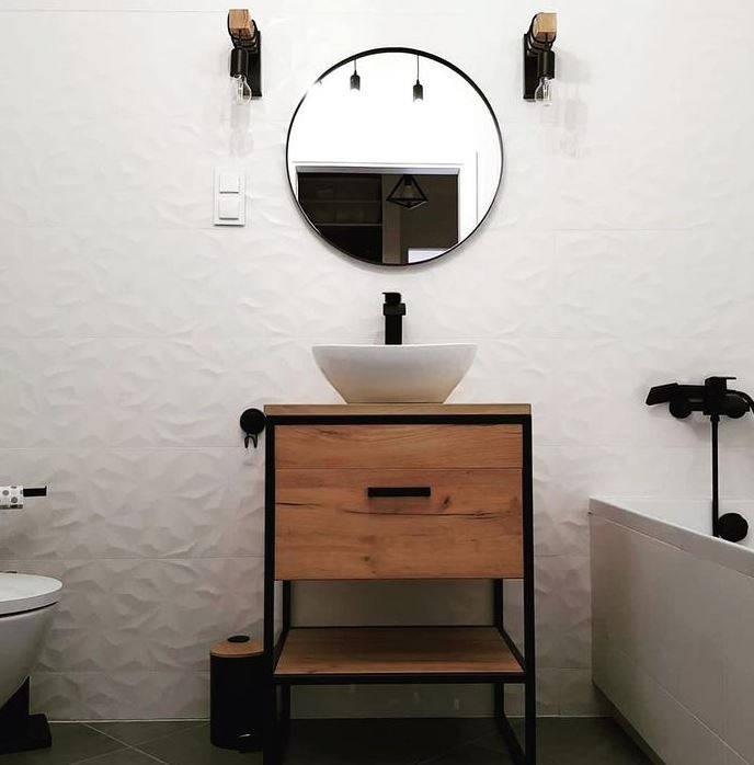 Nowoczesna łazienka z umywalką nablatową w kształcie misy, drewnianą szafką z czarną ramą, okrągłym lustrem i białymi płytkami 3D na ścianie.