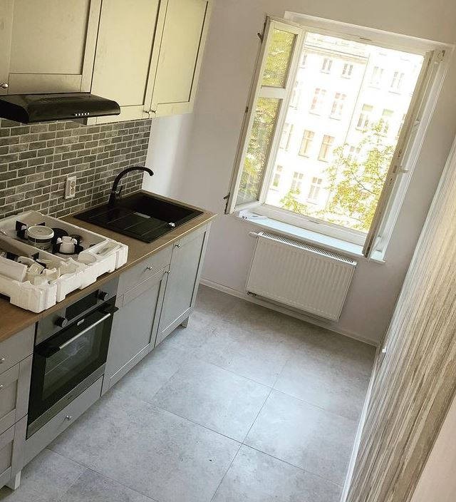 Nowoczesna kuchnia w trakcie montażu: szare szafki, czarny zlew, kuchenka z piekarnikiem w opakowaniu, okno z widokiem na budynek.