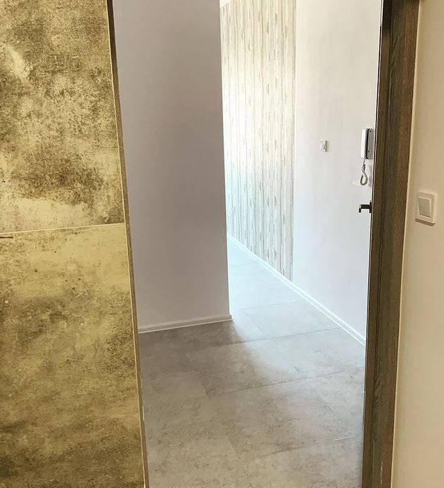 Korytarz w mieszkaniu z szarą podłogą z płytek, ściany w odcieniach bieli i beżu, widoczny domofon i włącznik światła, drzwi z drewnianą ościeżnicą.