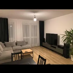 Jasne wnętrze salonu z szarą sofą narożną, drewnianym stolikiem kawowym, czarnym stołem jadalnym i krzesłami. Telewizor na niskiej szafce RTV, wysoka roślina doniczkowa w rogu. Okno z białymi...