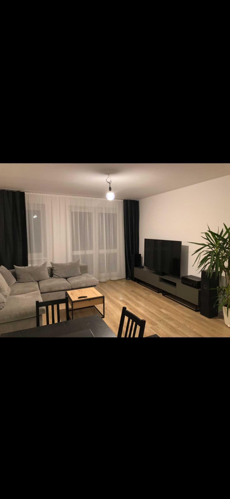 Jasne wnętrze salonu z szarą sofą narożną, drewnianym stolikiem kawowym, czarnym stołem jadalnym i krzesłami. Telewizor na niskiej szafce RTV, wysoka roślina doniczkowa w rogu. Okno z białymi...