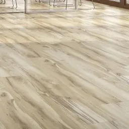 Jasne panele podłogowe z wyraźnym rysunkiem drewna, ułożone diagonalnie w przestronnym wnętrzu, z widocznymi nogami krzeseł i oknem w tle.