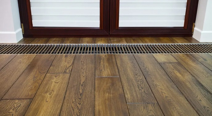 Parkiet z ciemnego drewna ułożony wzdłuż okna z żaluzjami, widoczna kratka wentylacyjna w podłodze.