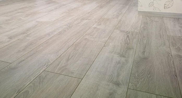 Jasnoszare panele podłogowe ułożone wzdłuż pokoju, widoczne fugi i wzór imitujący drewno, przy ścianie listwa przypodłogowa i fragment tapety w liście.