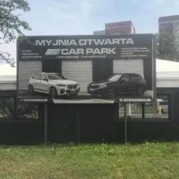 Billboard reklamowy myjni samochodowej Car Park z dwoma modelami BMW na tle szarej blachy falistej, z widocznym adresem strony internetowej i numerem telefonu, umieszczony nad budynkiem myjni...