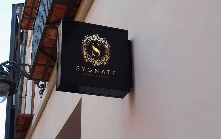 Czarny szyld z złotym logo firmy SYGNATE Angelika Sergot, zamontowany na jasnej ścianie budynku, widoczny z dołu pod kątem.