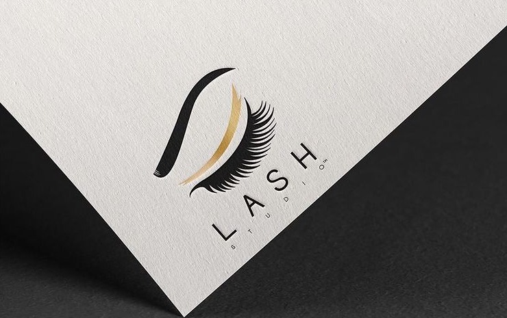 Eleganckie logo Lash Studio na teksturowanym papierze, przedstawiające stylizowane oko z czarną brwią, złotą powieką i czarnymi rzęsami, umieszczone ukośnie na białym tle z czarnym cieniem.