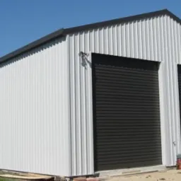 Gotowa hala z białej blachy trapezowej z dwoma czarnymi bramami rolowanymi i małym oknem po lewej stronie, na tle błękitnego nieba.