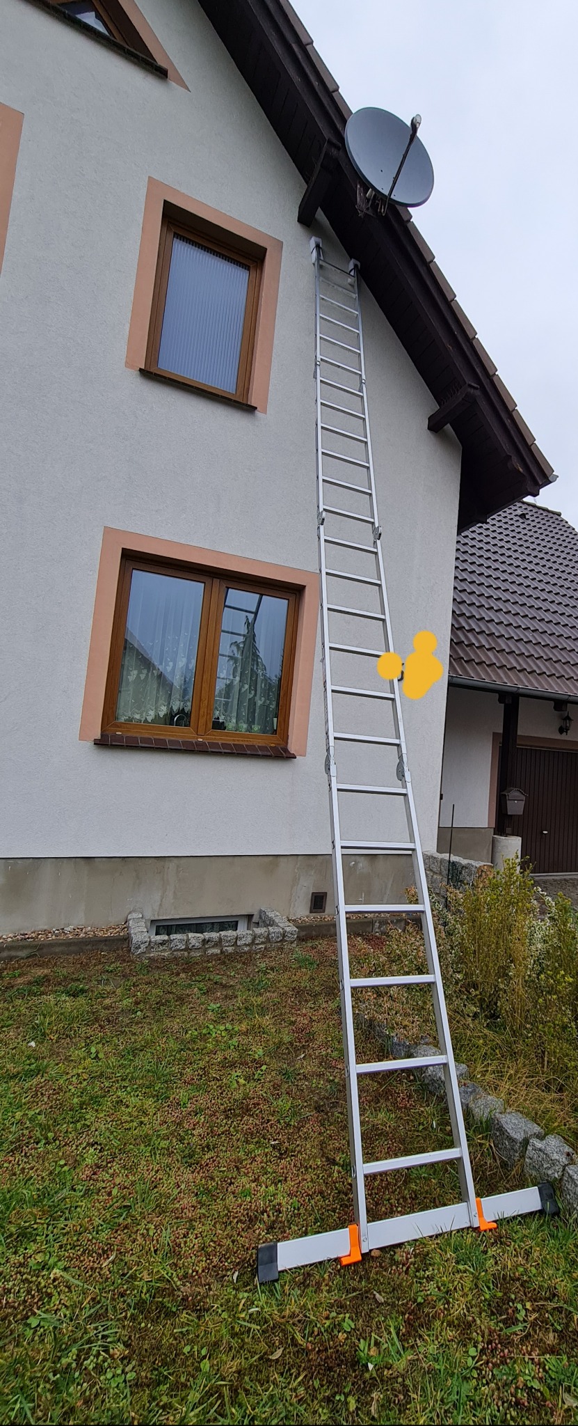 Aluminiowa drabina oparta o ścianę domu z zamontowaną anteną satelitarną. Zwracają uwagę pomarańczowe obramowania okien i podstawa drabiny z szerokimi, pomarańczowymi elementami stabilizującymi.