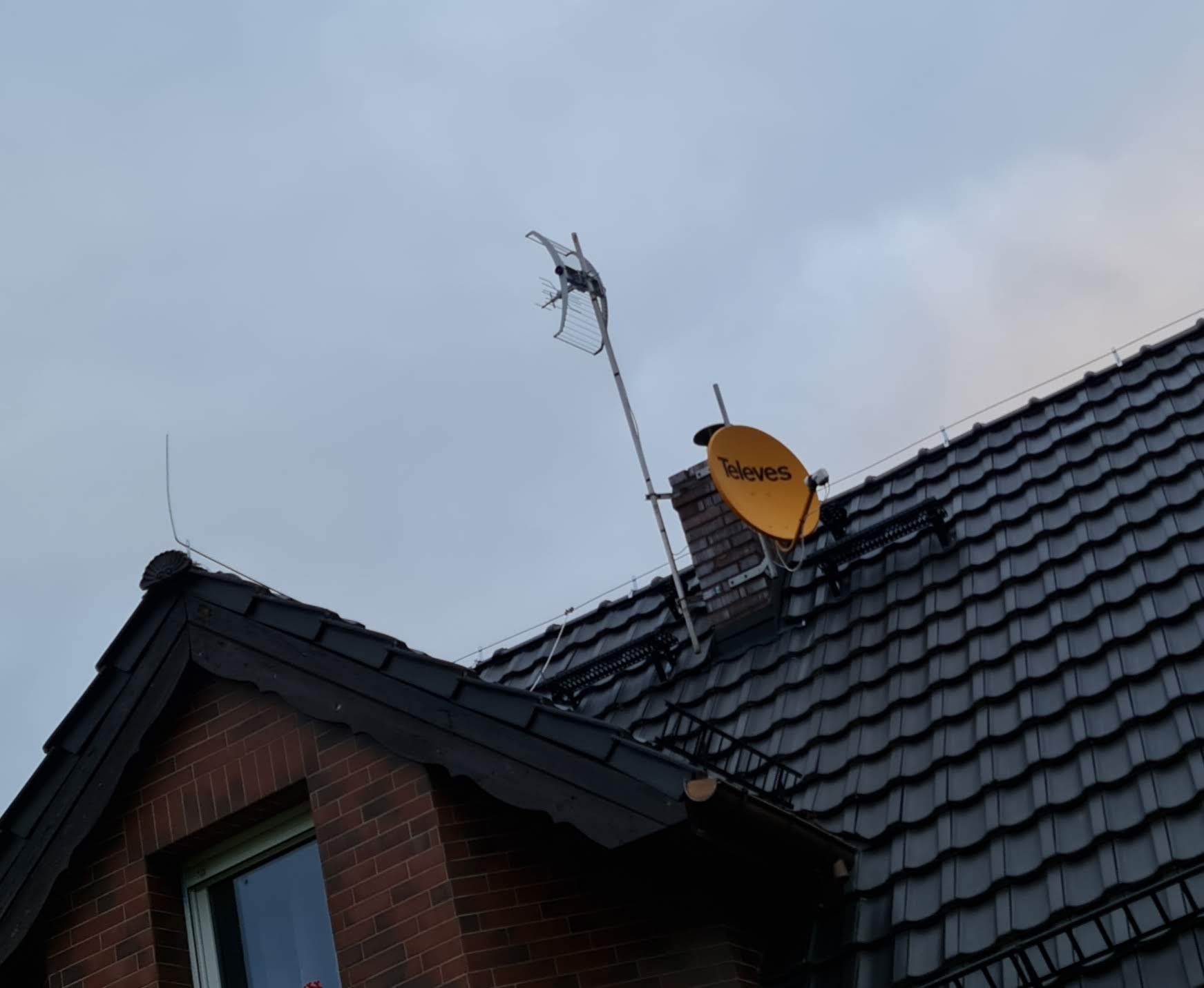 Antena telewizyjna Televes i antena kierunkowa zamontowane na dachu domu z czarną dachówką, widok z dołu na tle pochmurnego nieba.