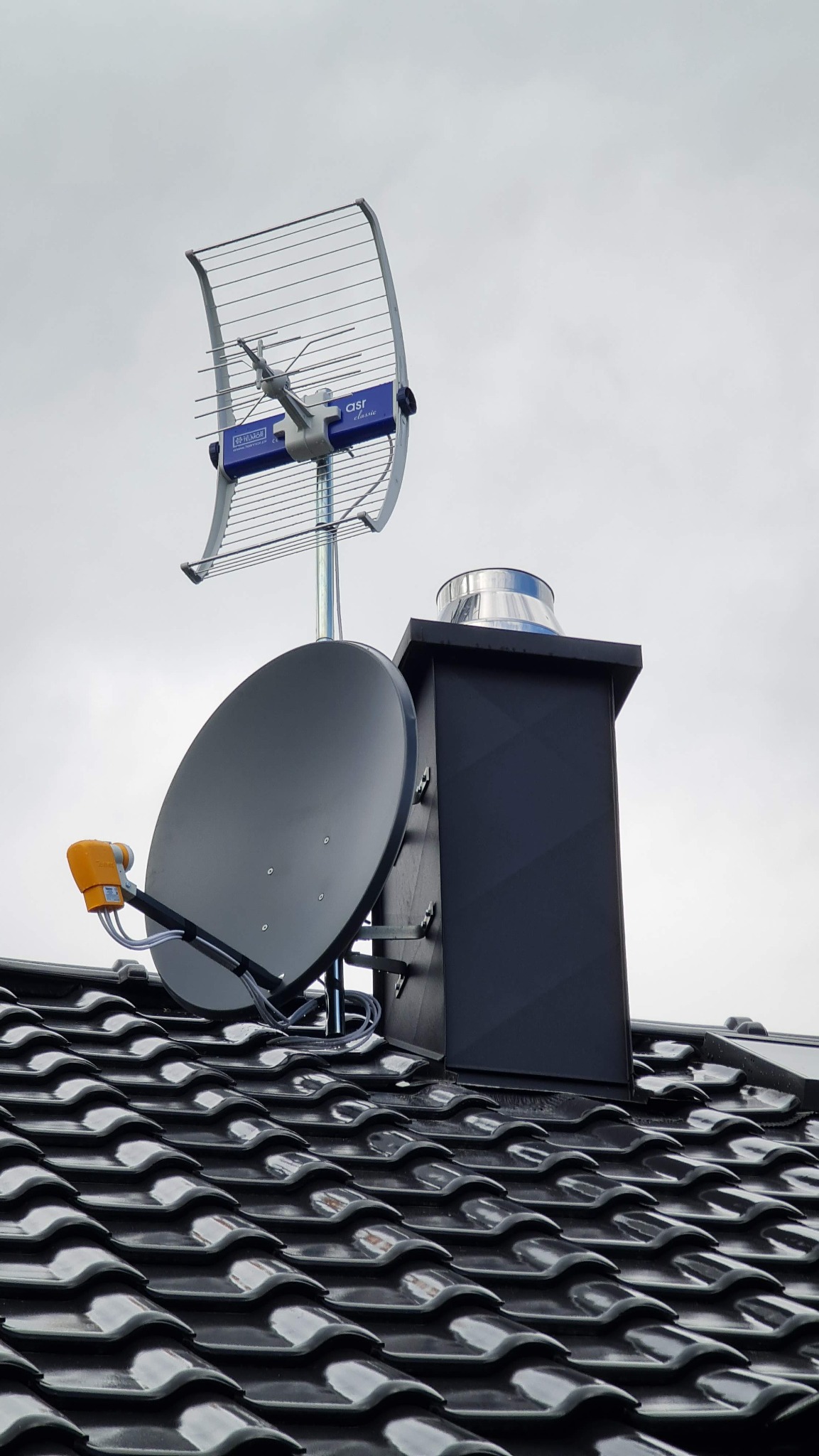 Antena Yagi-Uda i antena satelitarna zamontowane na dachu obok komina, widok z dołu na tle pochmurnego nieba.