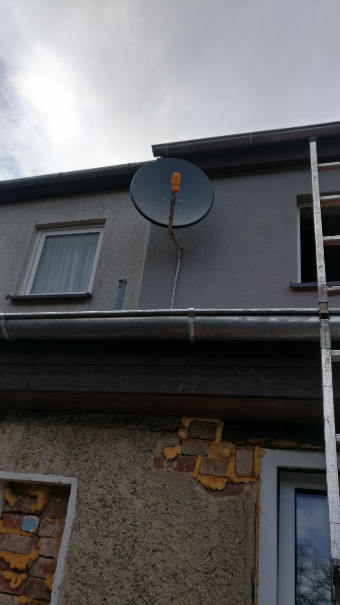 Czarna antena satelitarna z pomarańczowym elementem mocującym, zamontowana na szarej ścianie budynku, widoczna rynna i drabina oparta o ścianę.
