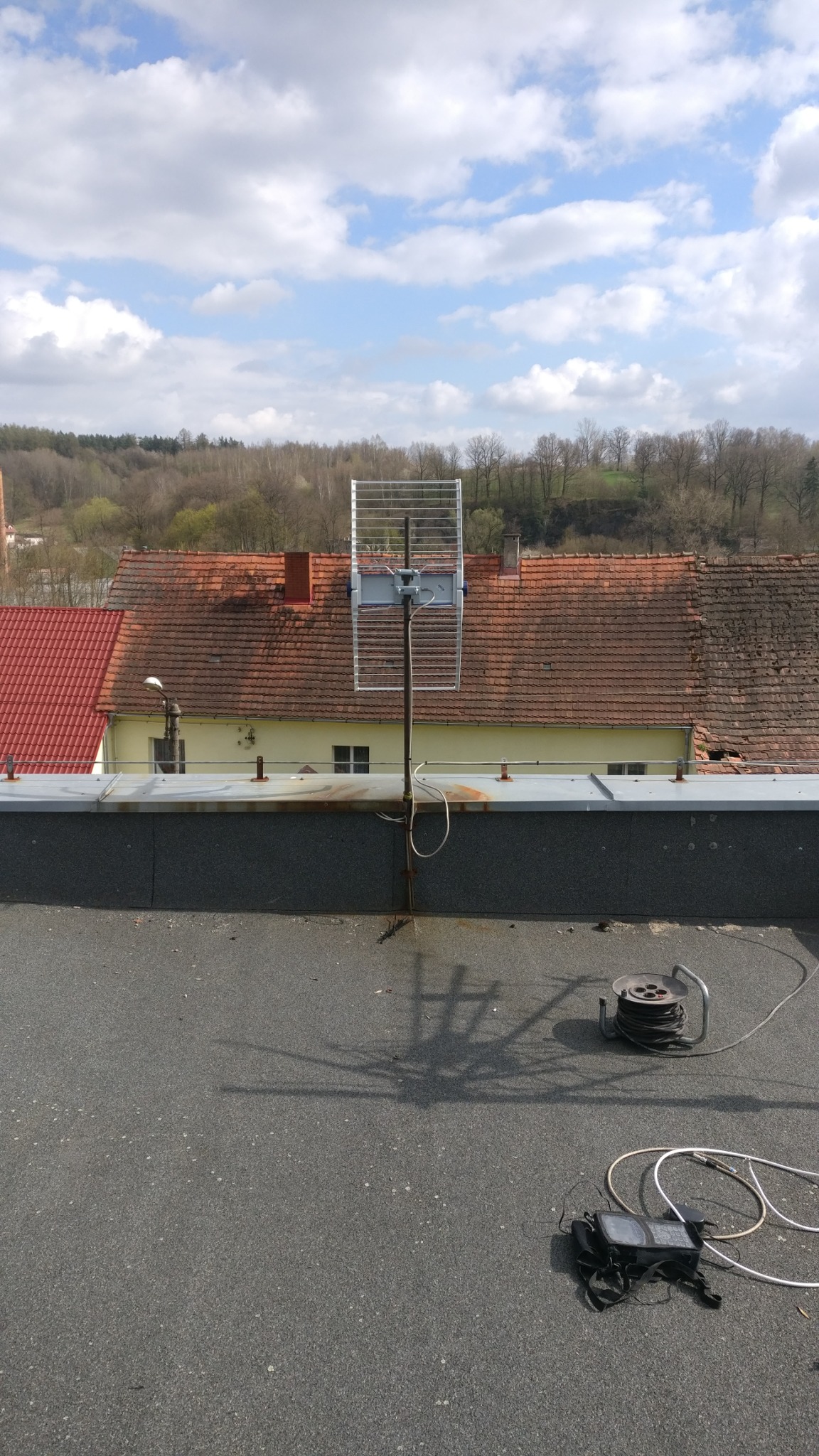 Antena siatkowa ustawiona na dachu budynku z widocznym kablem, miernikiem sygnału i zwiniętym przewodem. W tle zabudowa miejska i zielone wzgórza pod błękitnym niebem z chmurami.