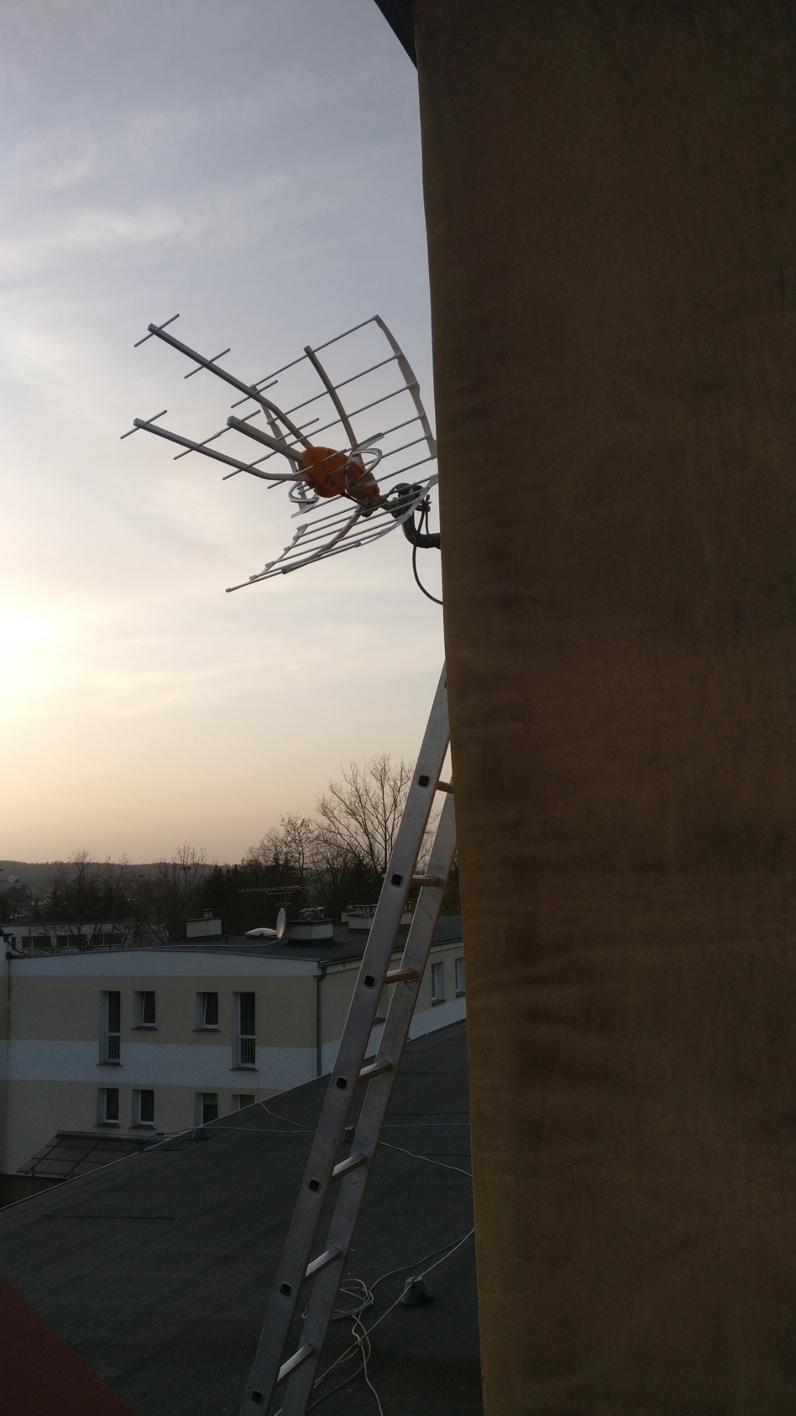 Antena telewizyjna zamontowana na dachu budynku, widoczna drabina oparta o komin, tło stanowi zabudowa miejska i zachodzące słońce.