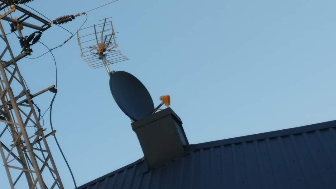 Antena satelitarna i antena telewizyjna zamontowane na kominie na dachu pokrytym blachą trapezową, widoczne linie wysokiego napięcia na tle błękitnego nieba.