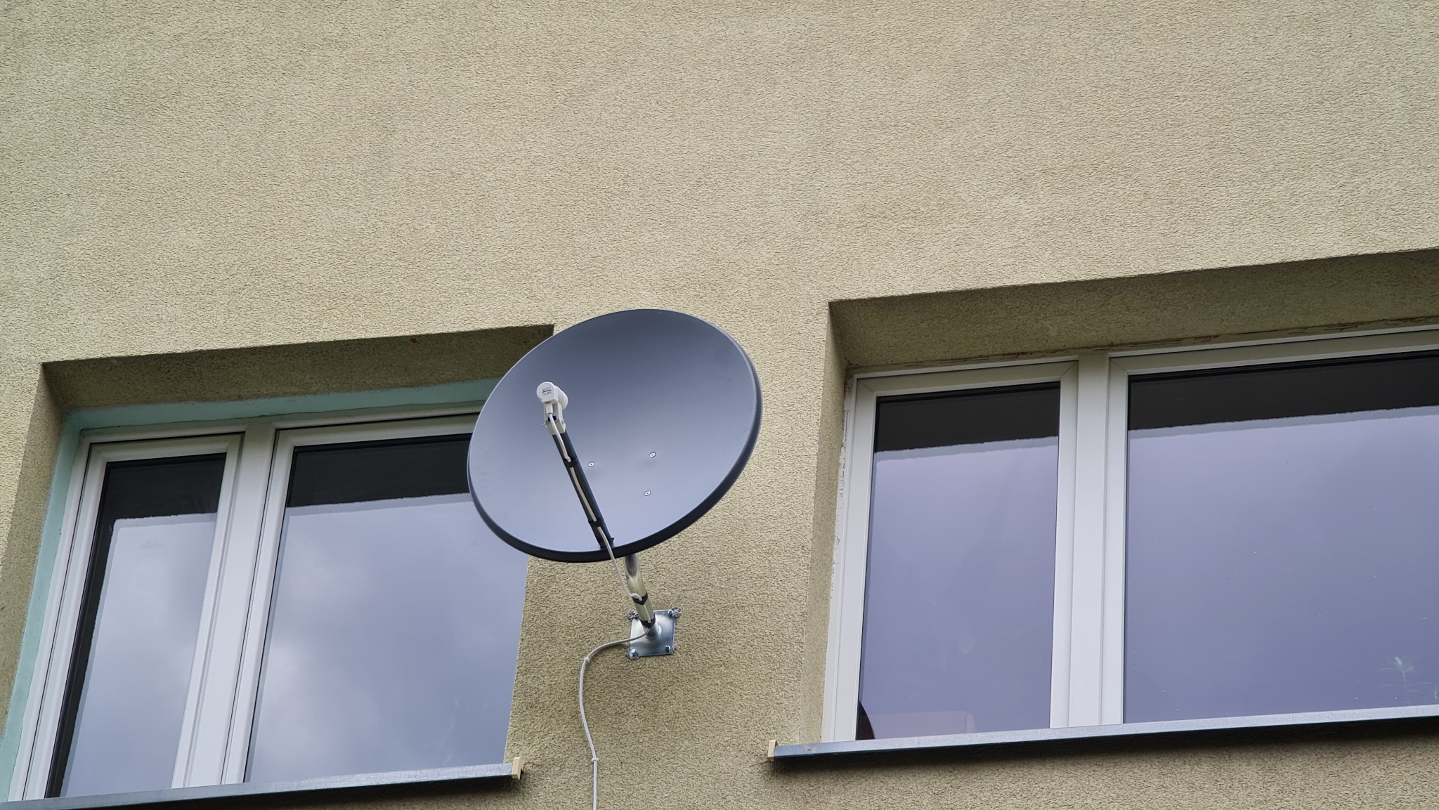 Antena satelitarna zamontowana na elewacji budynku, pomiędzy dwoma oknami z białymi ramami, widoczne okablowanie.
