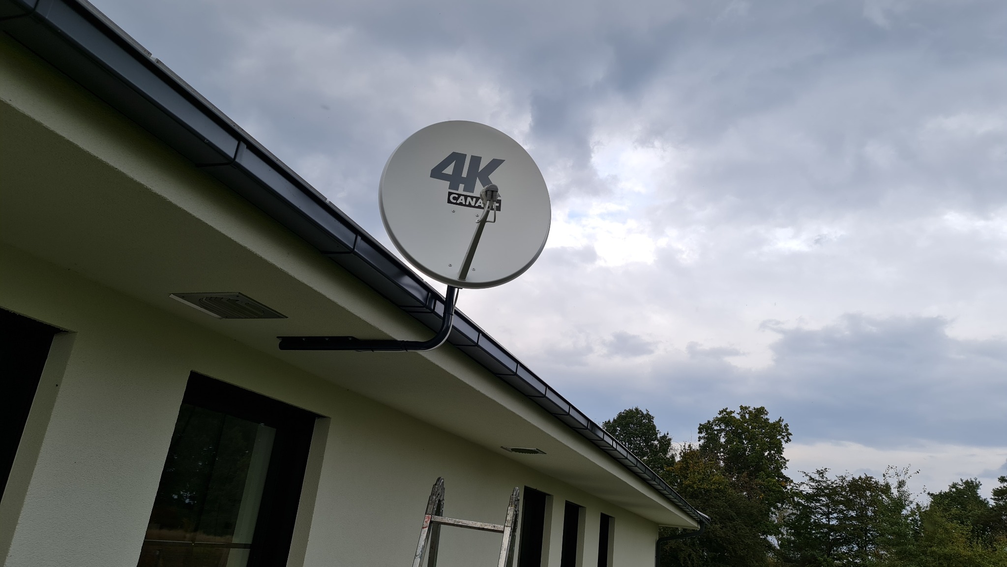 Biała antena satelitarna 4K Canal+ zamontowana na ścianie domu z widoczną drabinką i pochmurnym niebem w tle.