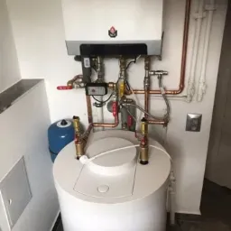 Kompaktowy zestaw grzewczy: biały zasobnik ciepłej wody i wiszący kocioł gazowy z miedzianą instalacją, widoczne zawory i czujniki, dodatkowy niebieski zbiornik wyrównawczy.