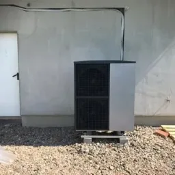 Zewnętrzna jednostka pompy ciepła, dwuczęściowa, zamontowana na betonowych bloczkach przed surową ścianą budynku z widocznymi przewodami i stertą cegieł w tle.
