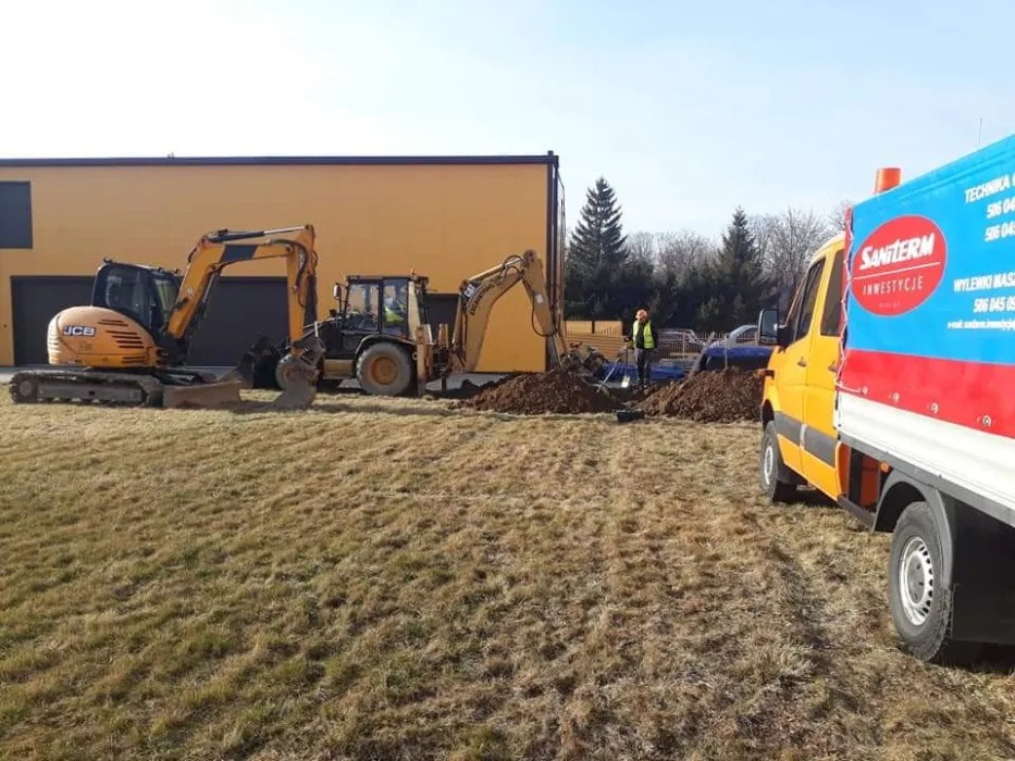 Dwie koparki JCB i CAT pracują przy wykopie na trawiastym terenie, widoczny pracownik w kamizelce odblaskowej i żółty samochód dostawczy z reklamą firmy Santerm Inwestycje, budynek w tle.