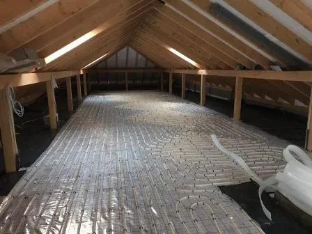 Instalacja systemu grzewczego podłogowego na poddaszu, widoczne rury rozprowadzone na czarnej folii izolacyjnej, drewniana konstrukcja dachu w tle.