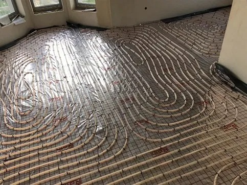 Rozłożone rury ogrzewania podłogowego na siatce montażowej, przygotowane do zalania wylewką, widoczne okna w tle i odbijające się światło słoneczne na folii izolacyjnej.