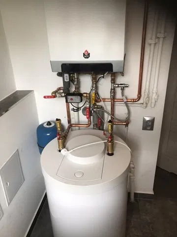 Kompaktowy zestaw grzewczy: biały zasobnik ciepłej wody i wiszący kocioł gazowy z miedzianą instalacją, widoczne zawory i czujniki, dodatkowy niebieski zbiornik wyrównawczy.