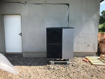 Zewnętrzna jednostka pompy ciepła, dwuczęściowa, zamontowana na betonowych bloczkach przed surową ścianą budynku z widocznymi przewodami i stertą cegieł w tle.