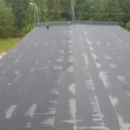 Widok z góry na czarny, bitumiczny dach po deszczu, z widocznymi śladami wilgoci, narzędziami dekarskimi i zielonym lasem w tle.