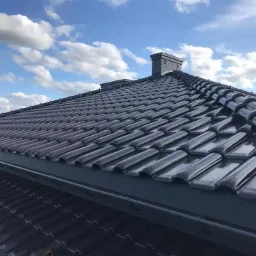 Błyszczący, ciemnoszary dach z dachówki ceramicznej, widoczny komin i rynna, na tle błękitne niebo z obłokami.