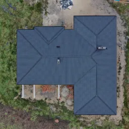 Widok z góry na ciemnoszary dach z blachy trapezowej na budynku w trakcie budowy, widoczne kominy, teren wokół częściowo porośnięty trawą, część placu budowy z porozrzucanymi materiałami.