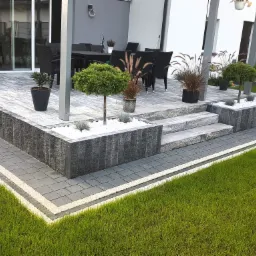 Nowoczesny taras z kostki brukowej w odcieniach szarości, schody kamienne prowadzące do części wypoczynkowej z meblami ogrodowymi, ozdobne rośliny w donicach i rabatach z białym kamieniem.