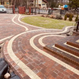 Kaskadowe schody i ścieżka z czerwonej kostki brukowej z jasnymi, piaskowymi akcentami, prowadząca przez trawnik w kierunku bramy wjazdowej na posesję z domem w tle.