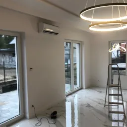 Wnętrze nowo wykończonego pokoju z zamontowaną klimatyzacją ścienną, widoczne okna balkonowe, drabina malarska i nowoczesna lampa sufitowa w kształcie pierścieni.