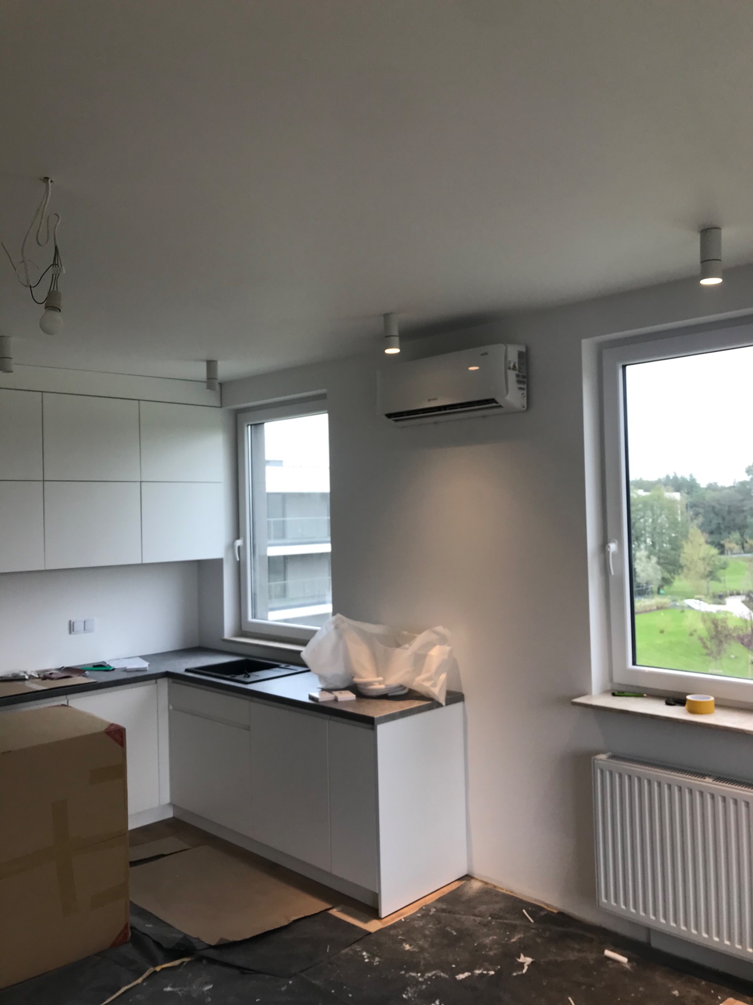 Wnętrze kuchni w trakcie remontu z zamontowaną klimatyzacją ścienną, widoczne zabezpieczenia podłogi, białe meble kuchenne i grzejnik pod oknem.