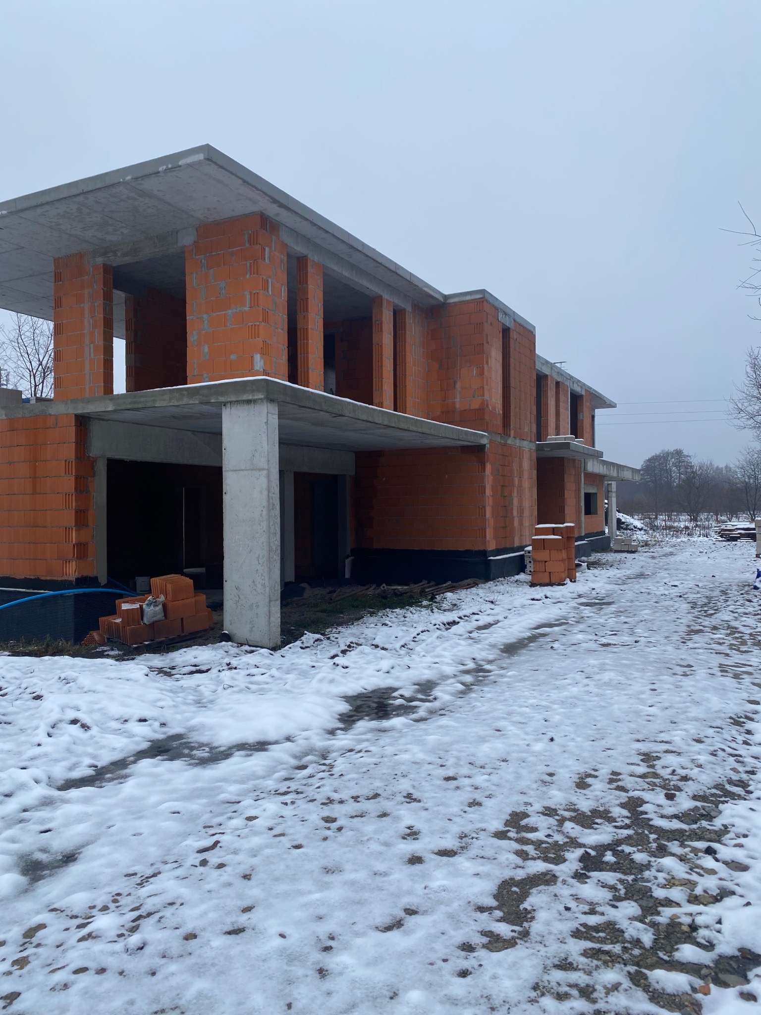 Surowy stan dwukondygnacyjnego domu z czerwonej cegły ceramicznej w zimowej scenerii, widoczne betonowe stropy i fundamenty, teren wokół pokryty śniegiem i błotem.
