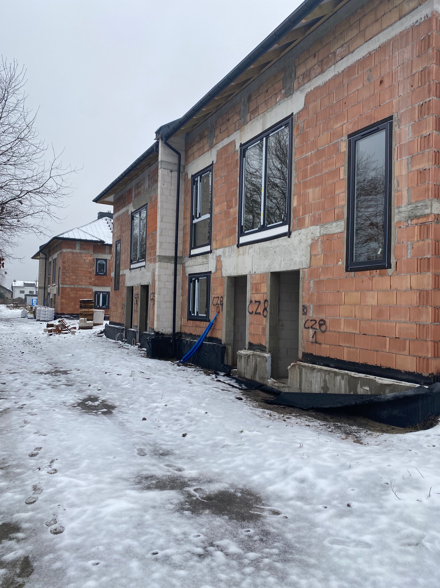 Surowe budynki w trakcie budowy, widoczne cegły, okna z czarnymi ramami, fundamenty i ośnieżone otoczenie.