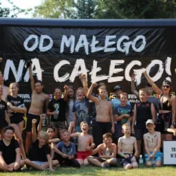 Grupa dzieci i dorosłych świętuje sukces przed czarnym banerem z napisem 'OD MAŁEGO NA CAŁEGO!' i tabliczką 'TRZEBA BYŁO ZOSTAĆ W DOMU I GRAĆ W GRĘ', wszyscy trzymają ręce w górze w geście triumfu.